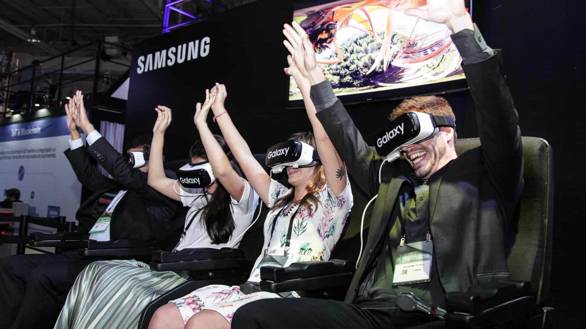 Pessoas rindo e se divertindo com o aparelho de Realidade Virtual da Samsung. Um homem está com as mãos para cima e gargalhando.